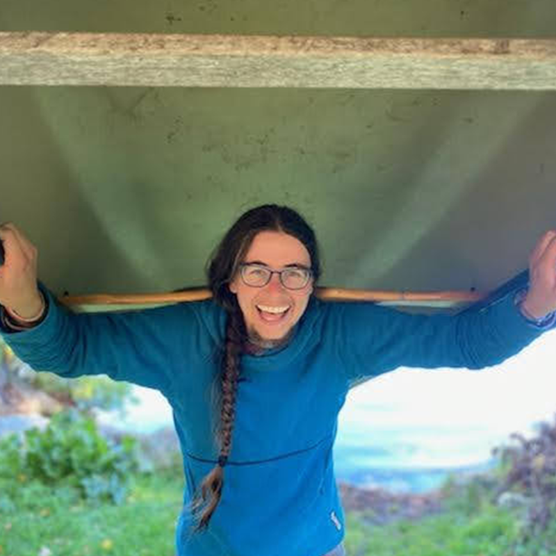 The image shows a person with long hair and glasses, smiling and holding a canoe over their head. They are wearing a blue sweater. The background appears to be outdoors, with some greenery visible. The person seems happy and is likely enjoying an outdoor activity.